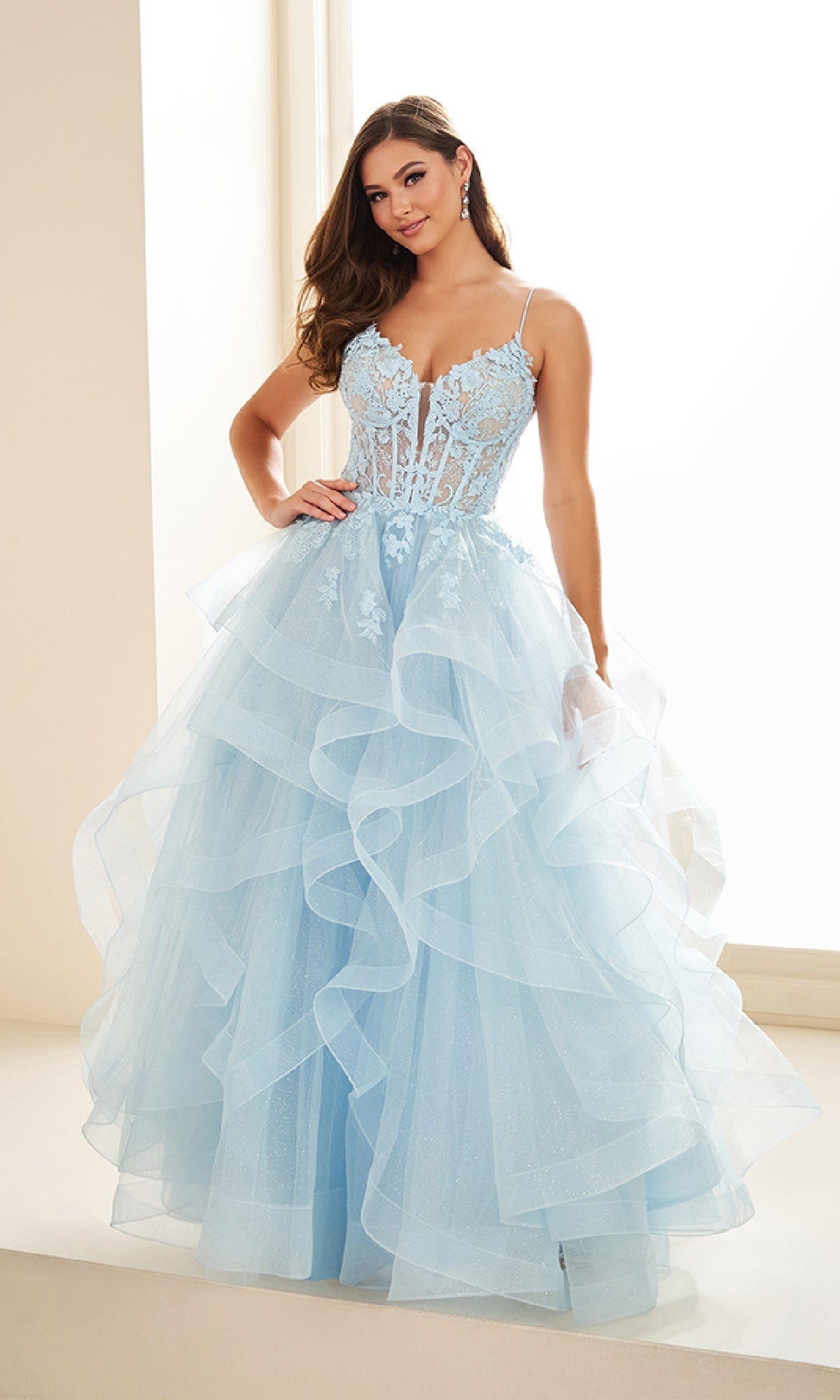 A woman stands indoors wearing the Ellie Wilde EW36203 Glitter Tulle Prom Ball Gown, which features floral lace detailing, an adjustable lace-up back, and thin spaghetti straps.